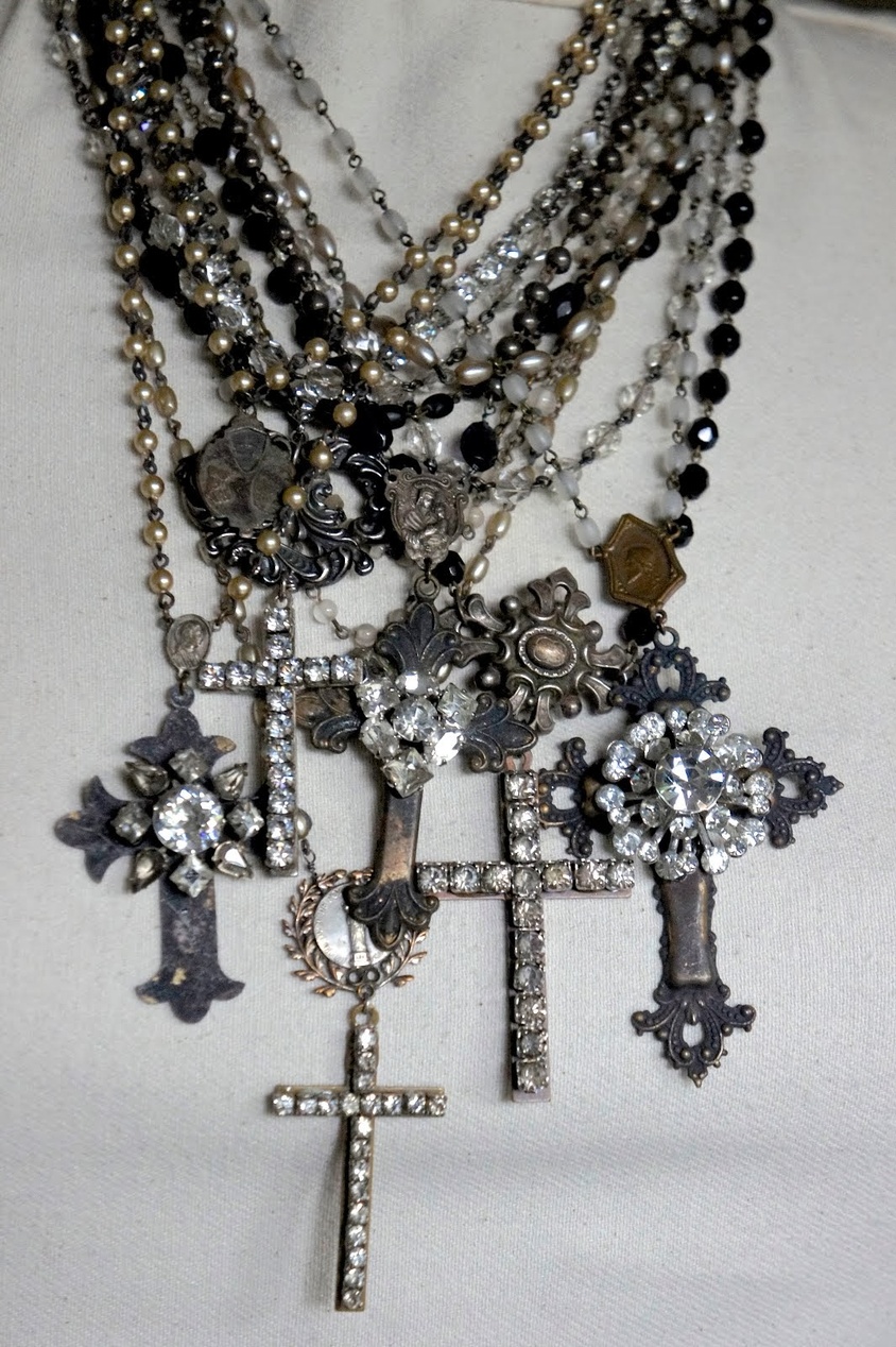 En samling lager-på-lager-halsband med olika korshängen, kristaller, pärlor och kulor i svarta, silver- och kristalltoner, visade på en vit tygbyst. Smyckena har en vintage, utsmyckad estetik.